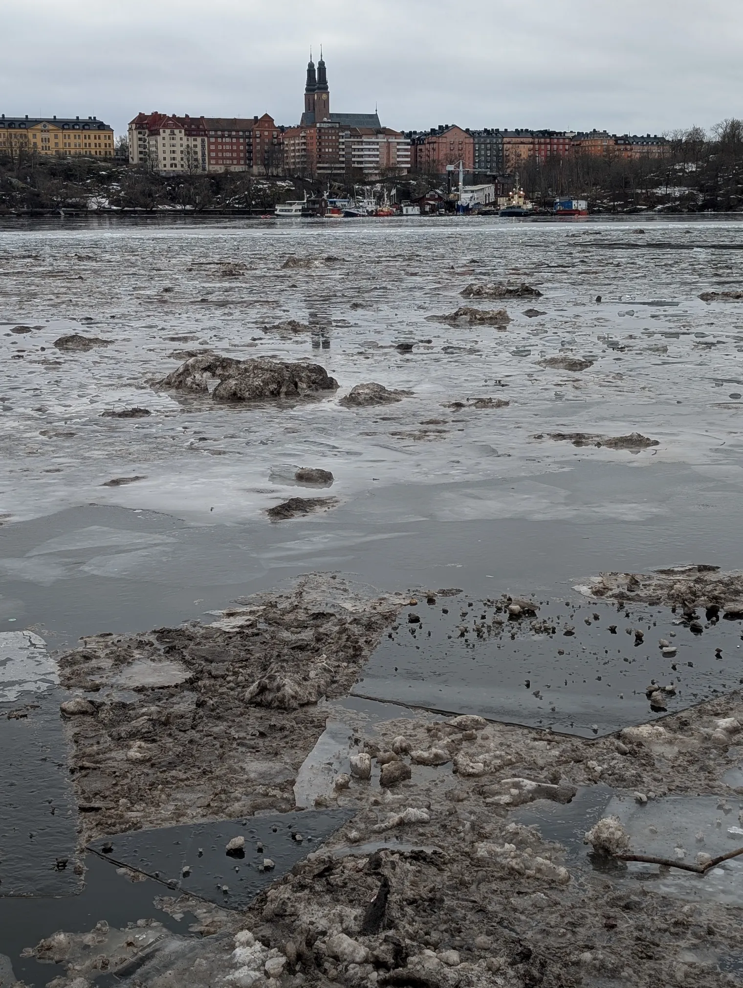 Ice floes covered in brown, dirty slush drifting on Riddarfjärden. Across the water, the Södermalm skyline is visible with the twin towers of Högalid Church.