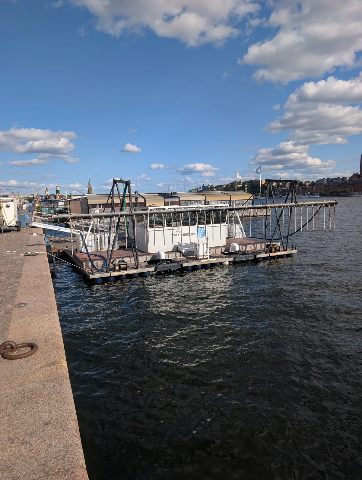 A white work pontoon marked Vattenresurs moored at a granite quay on a sunny summer day. A long boom with hanging hoses extends from the pontoon out over the water. Södermalm is visible across Riddarfjärden.