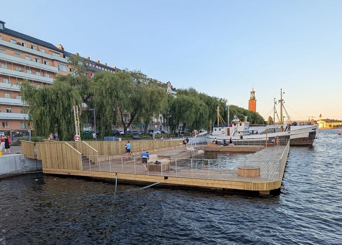 A newly built wooden pier with a swimming pool at the quayside on a summer evening. People are swimming and sunbathing on the pier, a white vessel is moored alongside, and the tower of Stockholm City Hall is visible in the background.