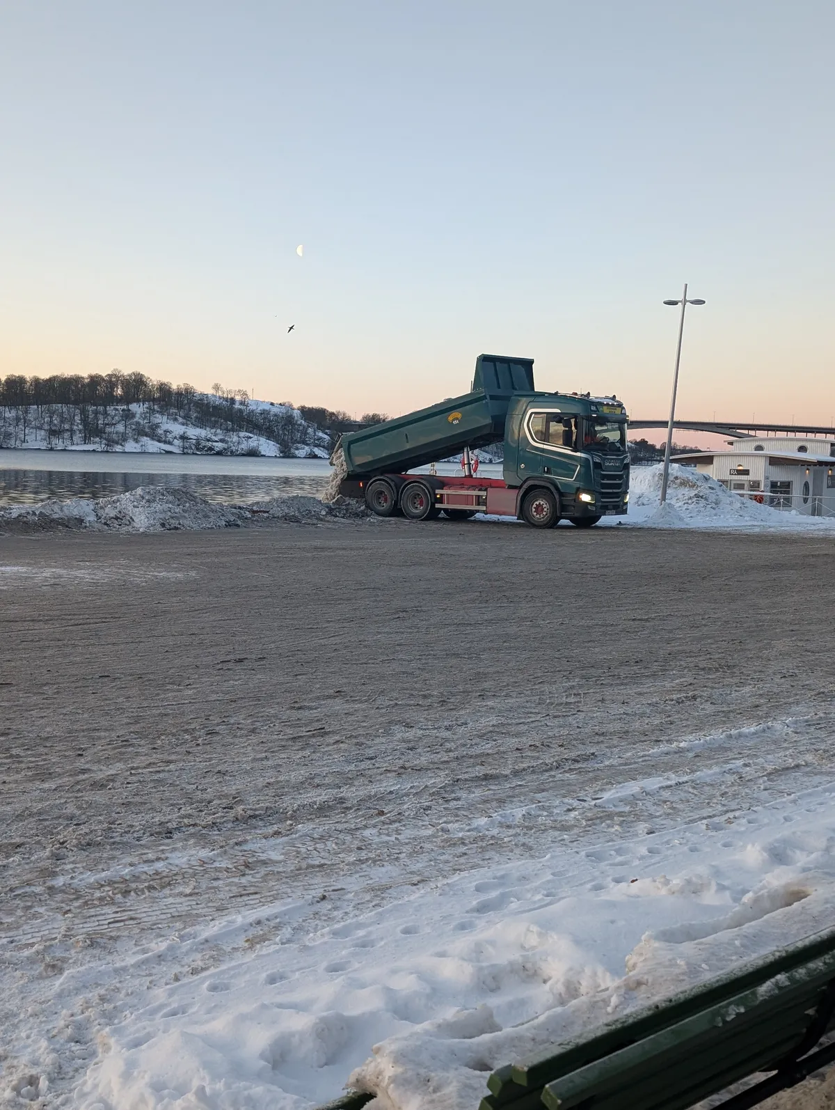 A green tipper truck discharging snow at the water's edge on a snow-covered quay at dusk. Lake Mälaren, a bridge and ploughed snow piles are visible behind the vehicle.