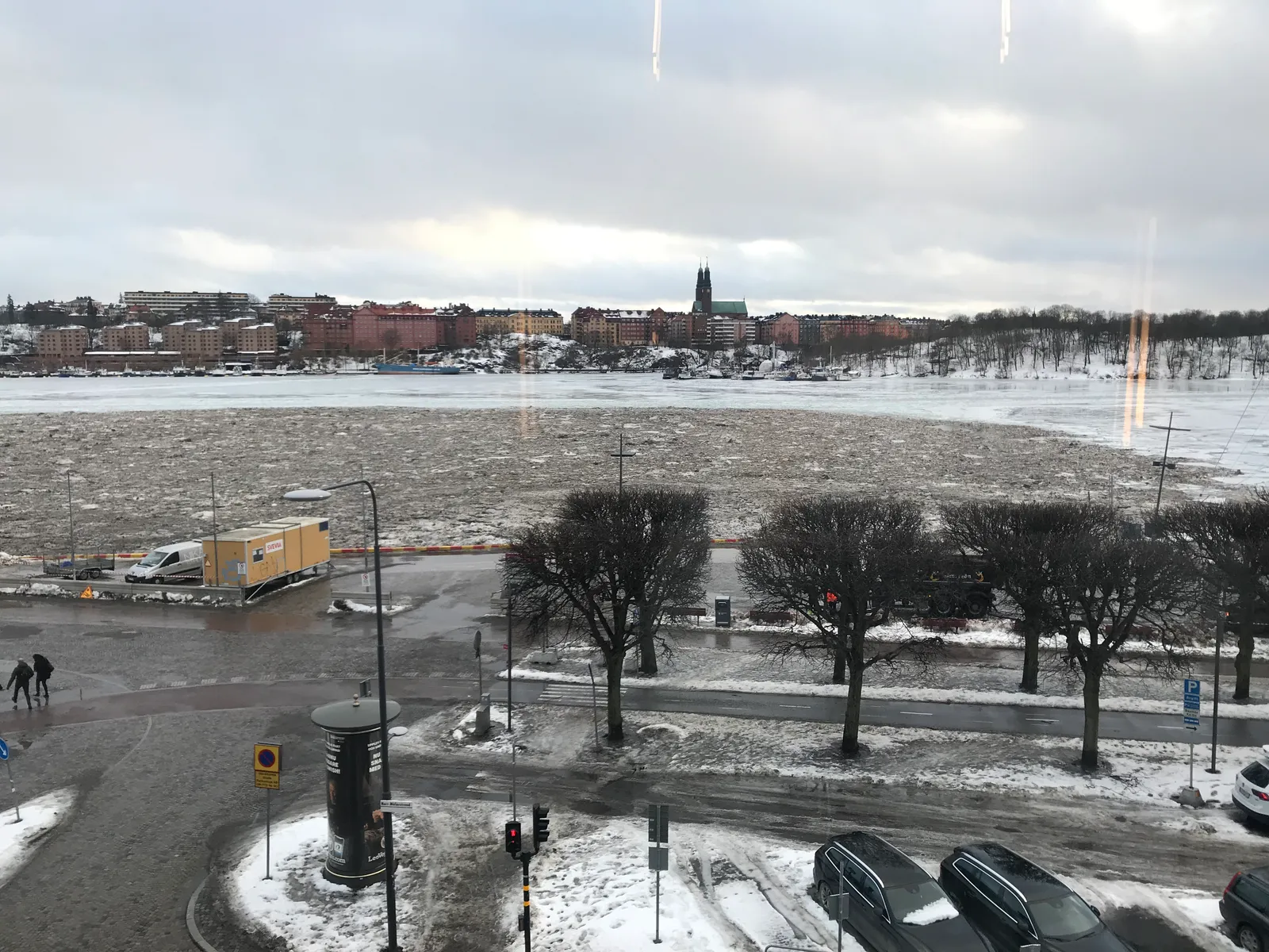 Riddarfjärden seen from above on a winter day. The water is covered in ice and brown, dirty slush, bare trees and parked cars line the quay, and across the water the Södermalm skyline is visible with the twin towers of Högalid Church.