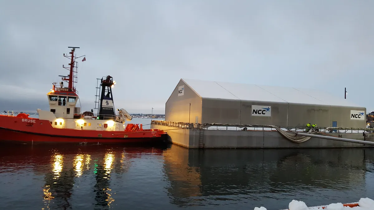 A snow melting plant on a barge moored at a quay at dusk, with the NCC logo on the white hall and a red tugboat alongside. Snow lies along the quay edge in the foreground.